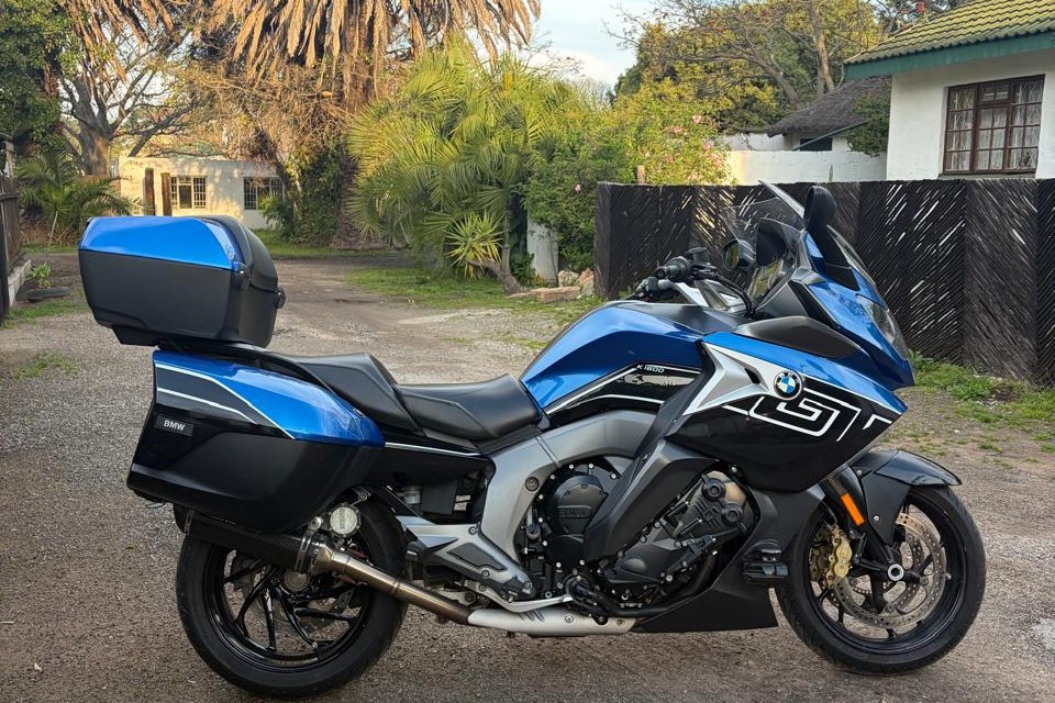 A blue BMW K1600 GT in full side profile, panniers and top box fitted, on a quiet lane framed by palms and gardens.